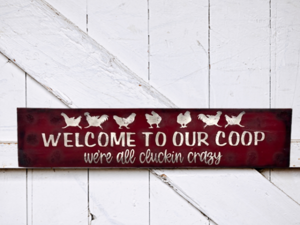 farmhouse chicken sign that has a deep red sawdust textured background with white chickens that reads Welcome to Our Coop, we're all cluckin crazy