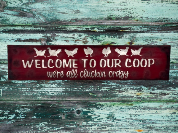 farmhouse chicken sign that has a deep red sawdust textured background with white chickens that reads Welcome to Our Coop, we're all cluckin crazy