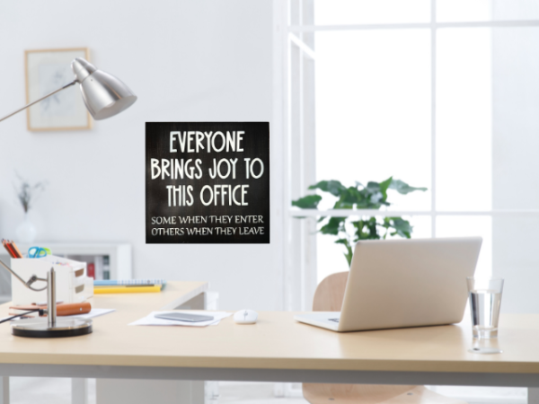 a humorous workspace sign with a dark brown background and cream colored letters that reads "Everyone Brings Joy To This Office - Some When They Enter, Others When They Leave