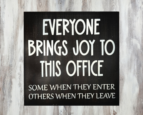 a humorous workspace sign with a dark brown background and cream colored letters that reads "Everyone Brings Joy To This Office - Some When They Enter, Others When They Leave