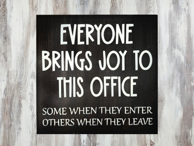 a humorous workspace sign with a dark brown background and cream colored letters that reads "Everyone Brings Joy To This Office - Some When They Enter, Others When They Leave