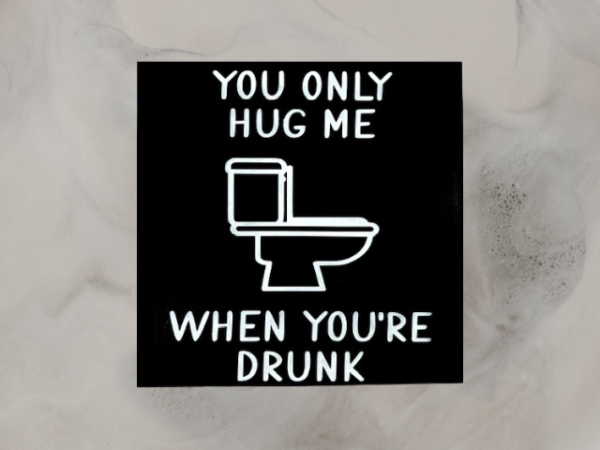 humorous bathroom sign with a black background and white wording that reads "You Only Hug Me When You're Drunk" featuring a toilet in the middle of the sign