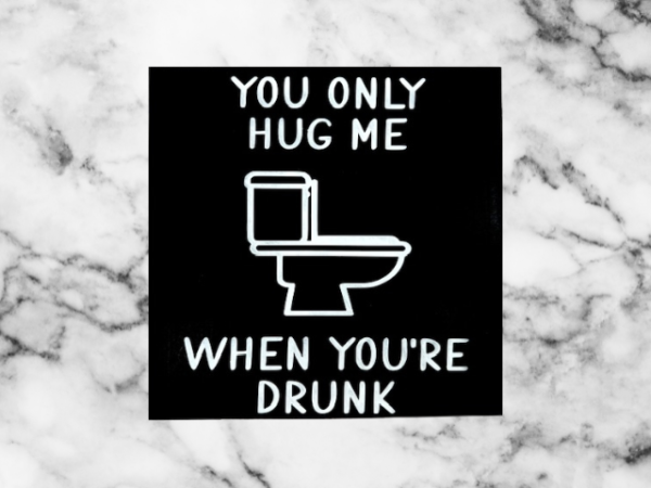 humorous bathroom sign with a black background and white wording that reads "You Only Hug Me When You're Drunk" featuring a toilet in the middle of the sign