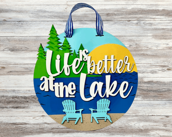Laser-cut wood lake house decor sign in vibrant colors that reads "Life is Better at the Lake", featuring trees, sun, and beach chairs on a sandy beach