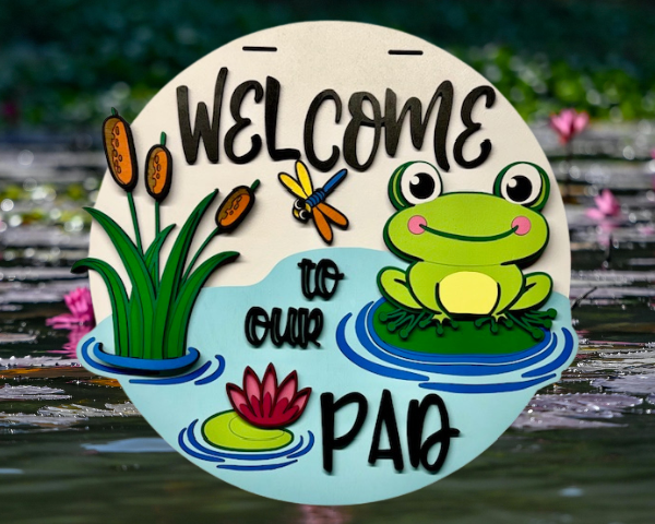 3D laser cut wood welcome sign with a neon green frog sitting in a pond surrounded by lily pads and grass, featuring the phrase "Welcome to Our Pad"